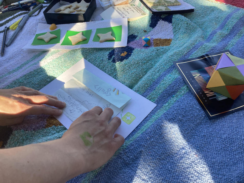 wingy's hands demonstrate an origami fold on a picnic blanket covered in snacks